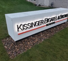 Illuminated sign on a stone base reads "Kissinger Bigatel & Brower REALTORS," located on a grassy area.