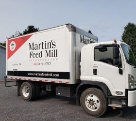 A white delivery truck with "Martin's Feed Mill" branding is parked on a gravel driveway next to a building.