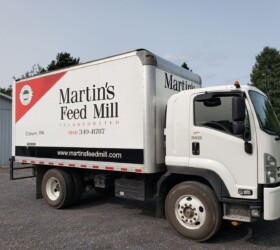 A white delivery truck with "Martin's Feed Mill" branding is parked on a gravel driveway next to a building.